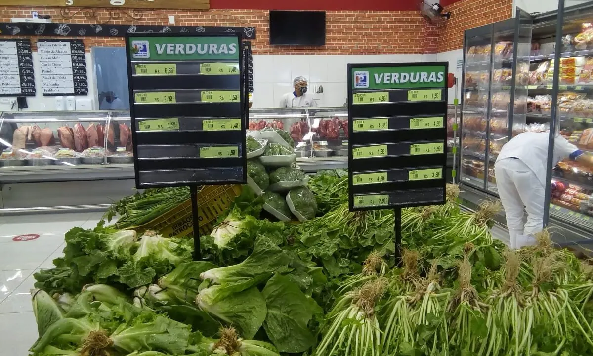 Duas placas pretas escritas Verduras, com linhas de preço, presas em hastes sobre uma banca de folhas verdes