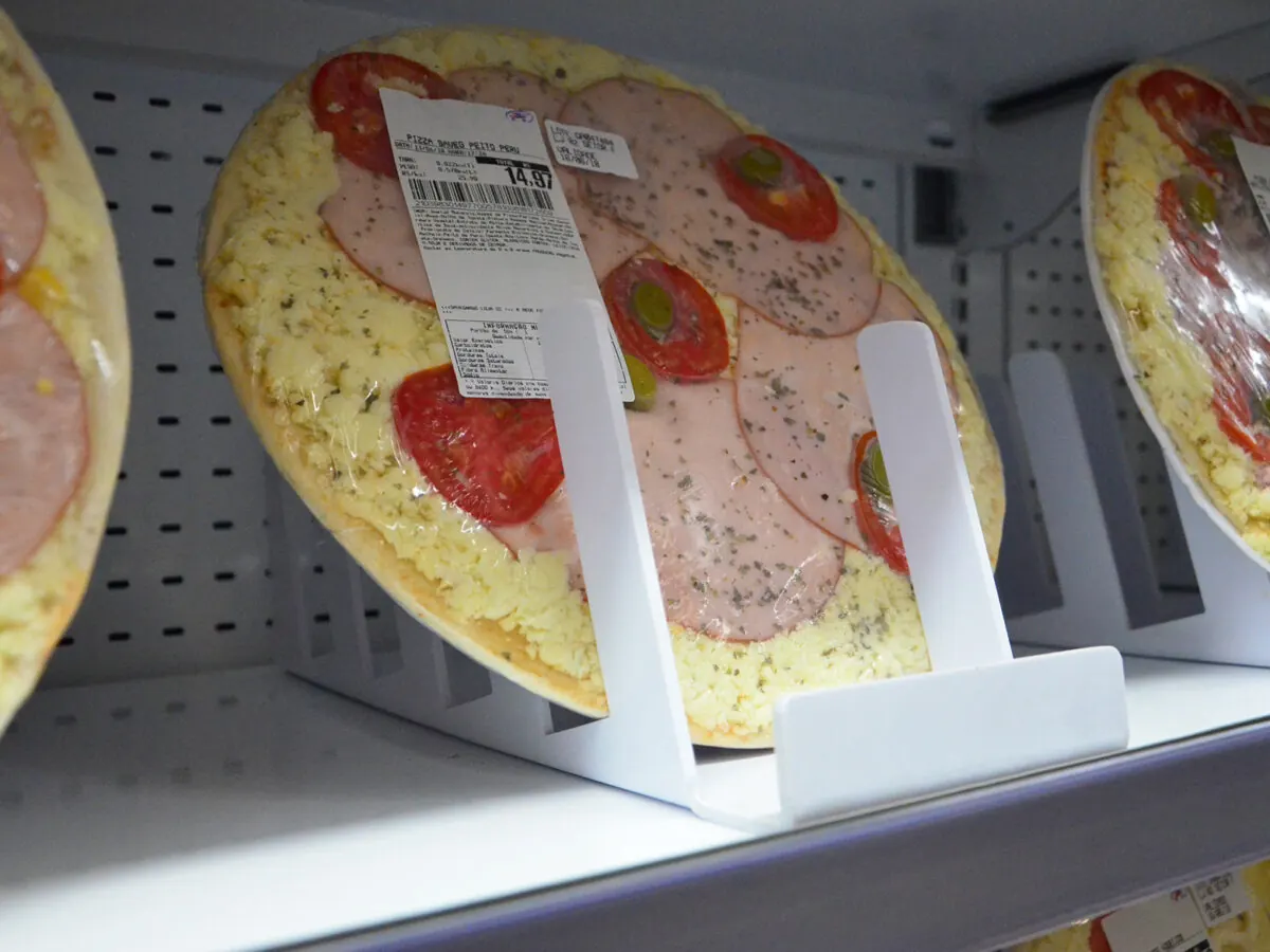 Pizza embalada em pé, apoiada em um suporte branco na prateleira refrigerada, com a etiqueta de preço do mercado à vista