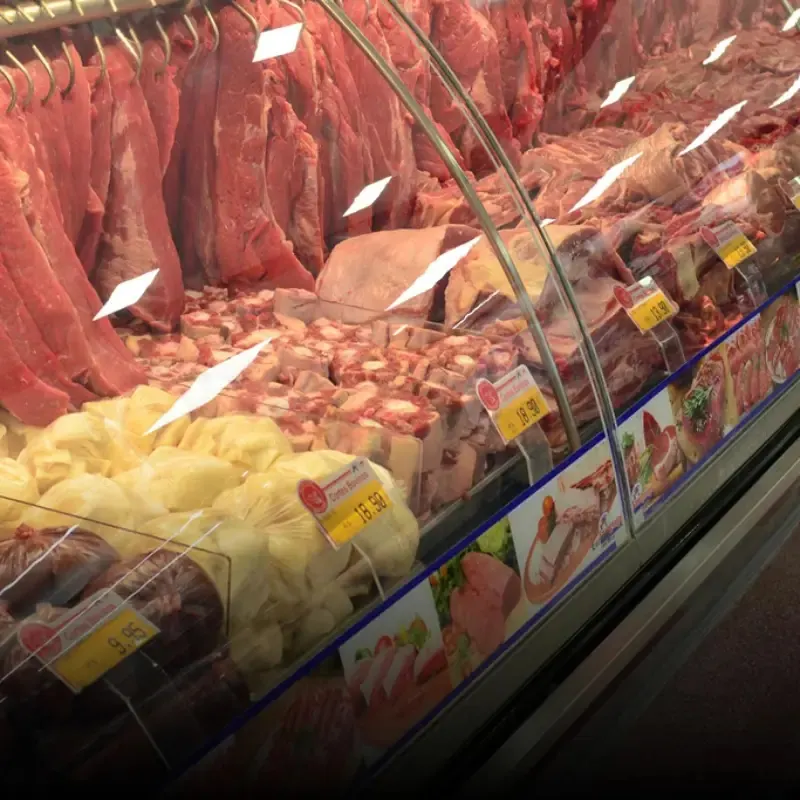 Balcão de açougue de vidro curvo com carnes expostas e etiquetas de preço em suportes transparentes entre os cortes